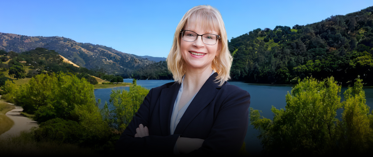 Attorney Kim Cameron-Smith standing with arms crossed in front of a scenic lake and hills